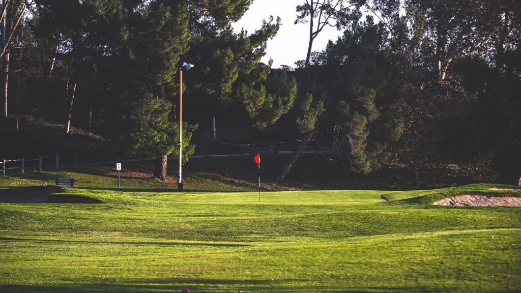 Putting green at Lake Forest Golf and Practice Center in Lake Forest, California, surrounded by mature trees and rolling turf under afternoon sunlight