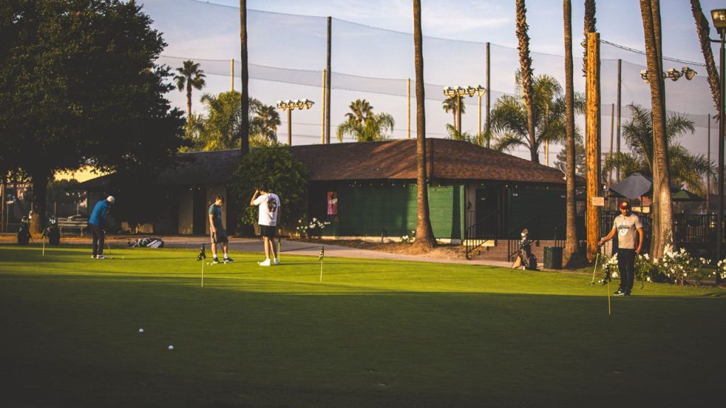 Golfers practicing putting at Lake Forest Golf and Practice Center, with palm trees, practice facility buildings, and netted driving range visible in the background.